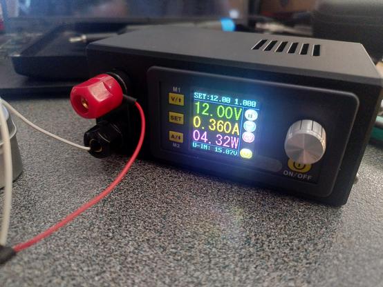 A small black bench power supply the screen on the front shows 12.00V, 0.360A, 04.32W. there's a red and white wire going off to the left. The background is blurry.