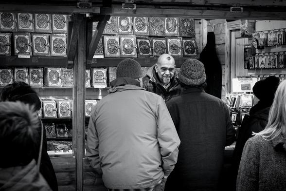 A man is working behind the counter of a craft stall at the Christmas Market at London's Trafalgar Square. He has a grey goatee beard and a small wisp of dark hair looking somewhat like a spider, or perhaps a small hairy Vesuvius on his shaved head.  He looks sad as he serves a man and a woman while others look at the goods on his stall. As well as fridge magnets and souvenirs, the main thing he's offering are leather-bound journals in three sizes, with some kind of decorative metalwork on the covers, some of which contains masks, faces, skeletal hands, anchors, ampersands, and elephants, along with many more abstract designs.