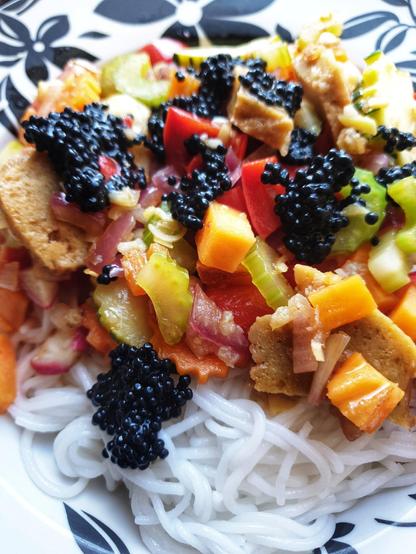 A bowl containing the meal. Stir fried spicy seitan and vegetables on top of rice noodles. Served with seaweed caviar on top of the stir fry
