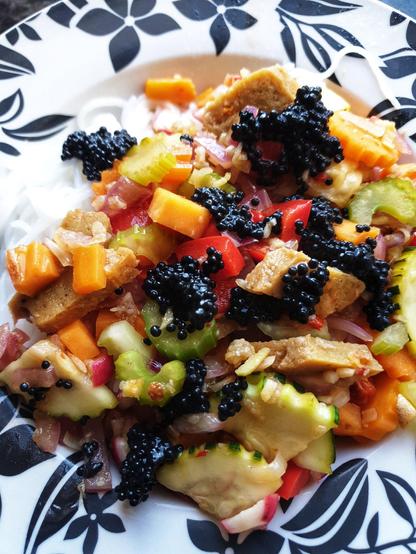 A bowl containing the meal. Stir fried spicy seitan and vegetables on top of rice noodles. Served with seaweed caviar on top of the stir fry