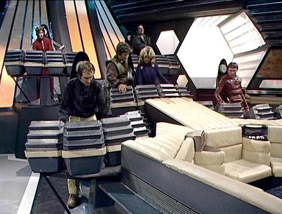 Claude 3.7 describes the image as: "This image appears to be from the classic science fiction television series "Blake's 7" from the late 1970s/early 1980s. The scene shows several crew members on the bridge or control room of their spacecraft, likely the Liberator.

The setting features the distinctive futuristic design elements typical of the show, with angular, geometric panels, hexagonal lighting fixtures, and the characteristic control stations where the crew members are positioned. There's a light-colored seating area or console visible in the foreground.

The crew members are wearing the distinctive costumes from the series, with some in darker attire and one figure visible in a reddish outfit. They appear to be at their stations on the bridge, which was a common setting for scenes in the show where the characters would plan their missions or navigate through space.

The image captures the retro sci-fi aesthetic that made this British television series memorable, with its distinctive set design and visual style that reflected the production values and creative approach of that era of science fiction television."

Blakes7Bot, version 3.9.24.