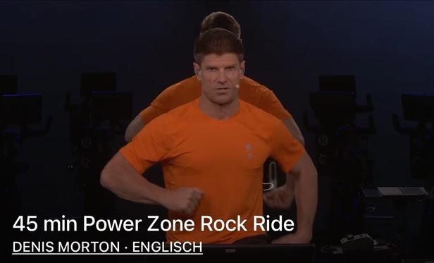 An instructor in an orange shirt is leading a 45-minute Power Zone Rock Ride workout. The background is dark, and exercise bikes are visible. Text at the bottom includes the instructor's name, Denis Morton