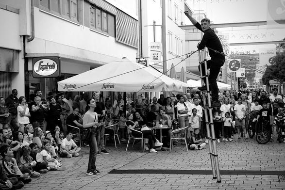 Black and white image of a street performer on a ladder in Esslingen, Germany. The performer, dressed in dark clothes, stands high on the ladder, arm raised. A crowd watches from below. Banners advertise a 'HOLZ OFEN PIZZA' and 'FESTIVAL'. Storefronts and onlookers fill the background, creating a lively street scene.