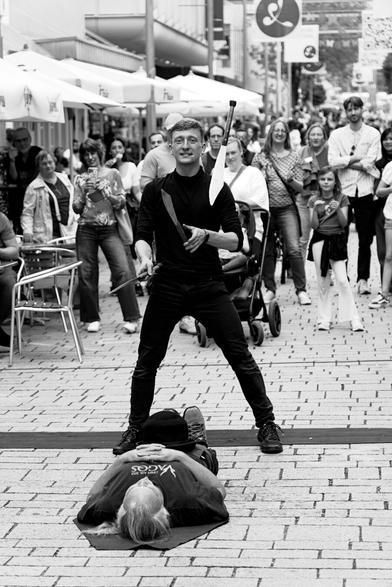 A black clothed man who is juggling knives over a man, who is lying on the ground. In the beackground many people are watching the scene