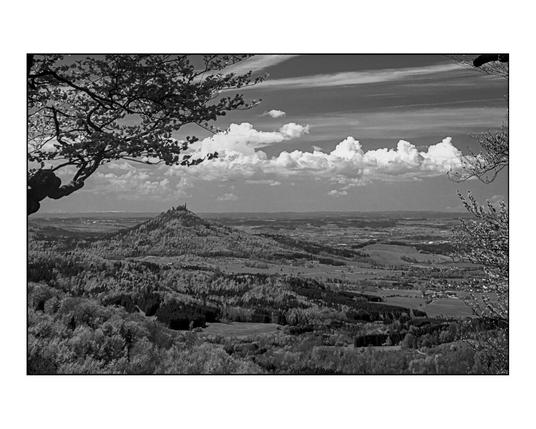 Black and white panoramic view of Hohenzollern Castle atop a prominent hill. The castle, with its towers and fortifications, stands silhouetted against a bright sky with scattered clouds. Rolling hills and valleys covered in forests stretch out in the foreground and middle ground, creating a sense of depth. Bare tree branches frame the upper left corner of the image. The sky features both fluffy cumulus clouds and streaks of cirrus clouds. The landscape appears vast and rural, with the castle as a central, imposing landmark.