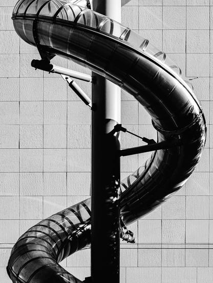 Black and white photo of a twisting metal tube slide mounted to a vertical pole, attached to a tiled wall. The slide’s transparent cover and reflective surface create dramatic light contrasts and geometric patterns.
