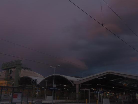 über dem Bahnhof haben sich dicke Regenwolken aufgestaut die noch leicht von der untergehenden Sonne auf der anderen Seite der Stadt mit einem Rest rot versehen sind und damit besonders bedrohlich erscheinen.