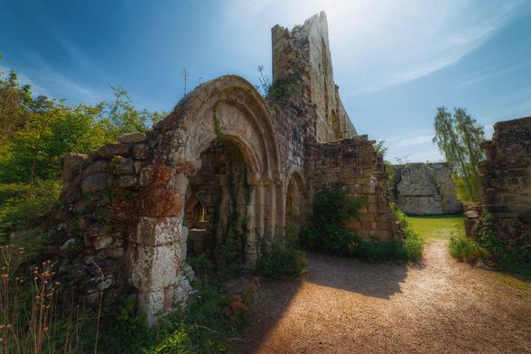 The sunlit ruins of Jervaulx Abbey in North Yorkshire on a clear day with a vibrant blue sky. The focal point is a detailed stone archway with weathered carvings, partially covered by creeping ivy and surrounded by lush greenery. Beyond the arch, further crumbling stone walls and arches frame a grassy courtyard bathed in warm sunlight. The scene captures the rich texture of the ancient stonework, overgrown plants, and a peaceful, almost timeless atmosphere, evoking the abbey’s historic significance and quiet beauty. Shadows cast by the ruins create depth and contrast on the dirt path leading through the site.