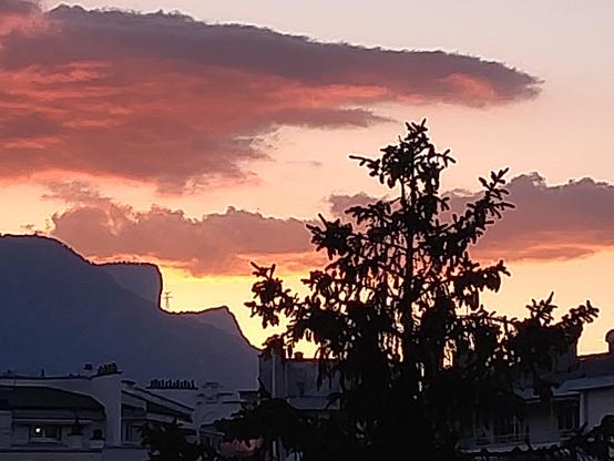 Nuages oranges du coucher de soleil au-dessus du bout du Vercors qui a une forme caractéristique en 2 marches d'escalier. Au premier plan toits d'immeubles dans l'ombre. Et un conifère qui se détache en noir sur fond orange.