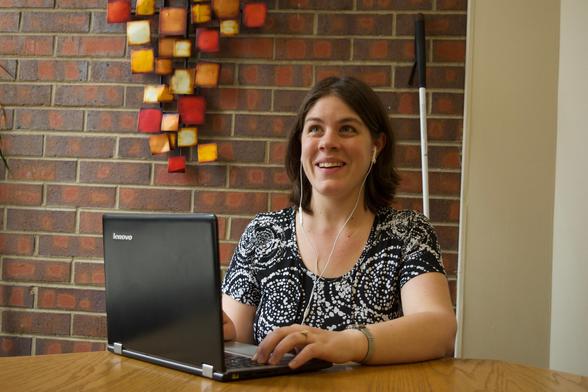Photo of a blind woman with earbuds, typing on her laptop while sitting by a round table.