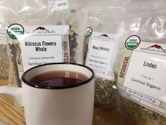 Photo of a teamug sat on a table. the tea inside is a rich red color. Behind are bags of bulk herbs stood upright: hibiscus flowers whole, wood betony leaf and flower, and linden leaf and flower.