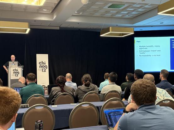 A presenter on stage with a slide on the screen that has the title “Pain” while describing intent and reality in automation