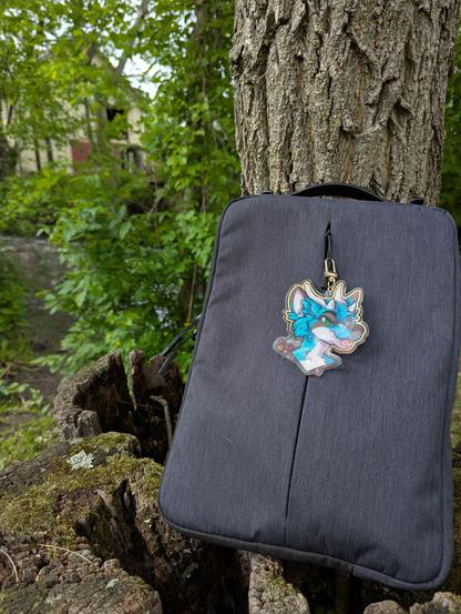 A laptop bag sitting on top of a hollow stump against a tree in front of a flowing river with an acrylic badge displaying a hellhound with teal fur and dark grey stripes, 4  curved horns, and emerald green eyes, sticking their tongue out