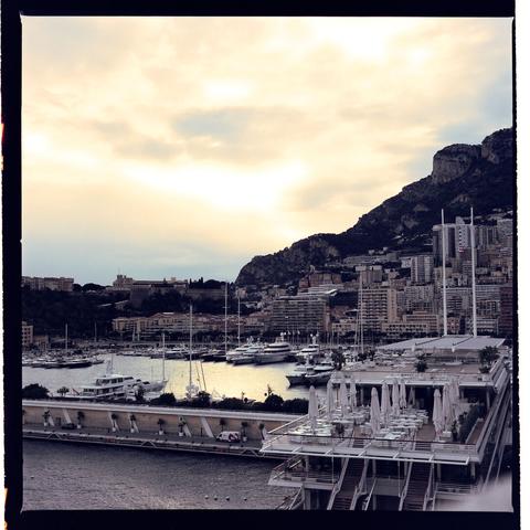 The image shows a coastal view with boats moored in a harbor, indicating a popular tourist destination. There are buildings along the waterfront, and mountains can be seen in the distance under an overcast sky during sunset.