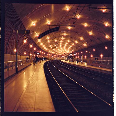 The image depicts an underground train station. There are railway tracks for trains to run on one side and pedestrian walkways on the other. Lights are installed along the ceiling of the tunnel-like structure, providing illumination for the station.