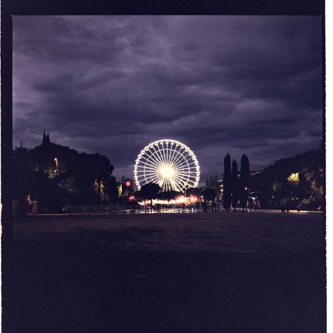 Image depicting an underground train station with lights along the ceiling, pedestrian walkways alongside railway tracks, and a large wheel structure in the background. Scene takes place at night under a cloudy sky.