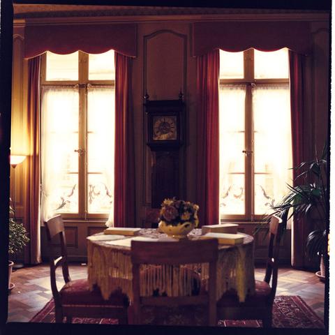 Indoor room at dusk with sunlight coming through uncurtained windows. Dining table set for four people, chairs on either side of the table. The room has wooden floors and appears to be a parlor or living room.