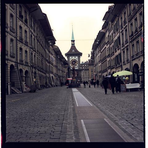 An empty narrow street with tall windows and wooden shutters. A covered walkway above the street connects buildings, hinting at the age of old town Bern.