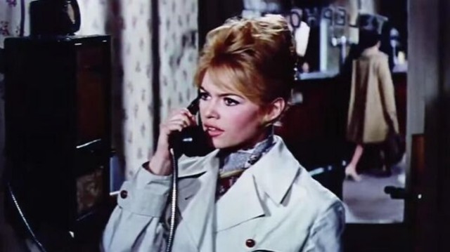In a still from a movie, Brigette is on the phone in an outdoor location.  She seems to be in a intense conversation.  She is wearing a light blue overcoat with a dark blue turtleneck underneath.  Her hair is up in a tight updo.