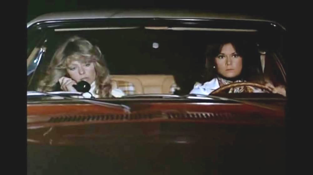 Farah Fawcett and Kate Jackson are seen through the windshield of a car.  Kate is driving and Farah is in the passenger seat of the car, with a standard phone receiver to her ear.  The car is red and it looks like the scene is taking place at night.