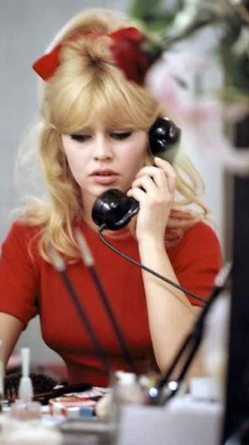 Brigette is in an perhaps an office setting on the phone.  She is shown from the waist up, sitting at a desk.  She is dressed in a red knit blouse.  Part of her hair is up with a big red bow, the rest falls down around her shoulders.