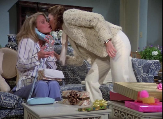 Cheryl Ladd and Jaclyn Smith are in an interior location.  Cheryl is sitting on a light colored couch and in front of her is a white table that has several wrapped presents that have colorful bows on them.  Also on the table is a blue princess phone and Cheryl has the receiver to her ear and has a glass vase in the other hand.  Cheryl is wearing a white jacket with blue stripes and blue slacks.  Jaclyn is leaning over in front of her wearing a off white wool sweater and white slacks.  The two women appear to be kissing each other.
