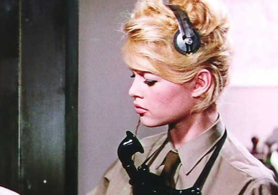 Brigette is shown as a either a radio or telephone operator in some sort of military setting.  She is wearing a khaki uniform shirt.  She has a headset on and a microphone on her chest that hangs from a  strap around her neck.  Her hair is in a tight updo.