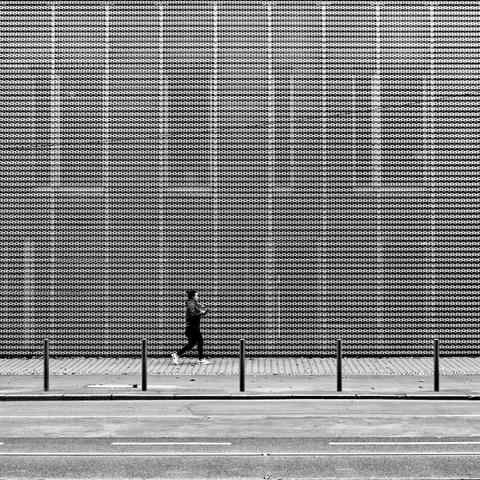 Black and white. square format. A passer-by walks past a façade, which can strain the eye when looking at it due to the perforated sheet metal cladding. Along the sidewalk are five posts that give the picture graphic order and structure.