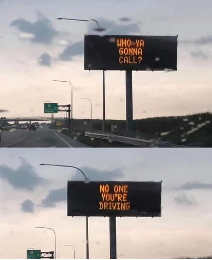 Digital road sign on a highway reads, “Who ya gonna call?” followed by, “No one. You’re driving.” Clouds and light rain in the background.