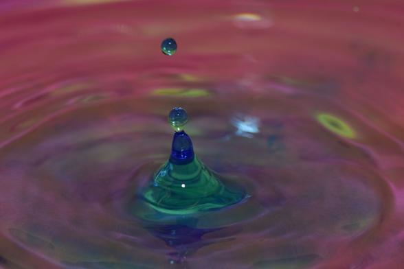 ein blau-grün aufsteigender Wasserkegel mit eiem Tropfen direkt darauf, sodass etwa die Form eines kleinen Männchens gebildet wird, und einem etwas darüber fallendem Tropfen vor pink-lila Hintergrund mit gelben Flecken.