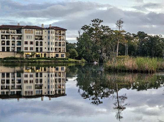 Colorful view across a quiet waterway. The opposite shoreline with a large luxurious building and the water winding through tropical foliage. The earthy colors of both cast a reflecting shadow upon the water below.