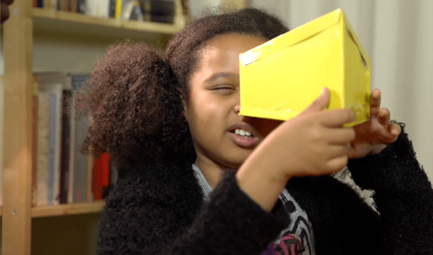 A young person with curly hair smiling while looking through a yellow cardboard box from one side, demonstrating the Ames Room illusion experiment from The Royal Institution's activity.