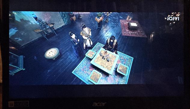 Bird's eye view of the male lead sitting at a table with four stools on a carpet. The ankle height braziers stands two meters away, behind his henchpersons. Another brazier has been placed smack on a carpet behind the male lead, close to a candle holder and a bonsai that stands in the middle of the floor. Other furniture are randomly strewn about, and the mass of just empty floor enhances the feeling of sterile interior design cluelessness.