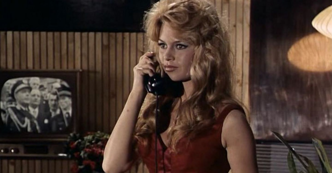 In a still from the movie "La Parisienne," Brigette Bardot is in an interior setting.  Behind her is a black and white television with an image of military people on the screen.  She is standing in the middle of the image, with a phone receiver up to her ear.  She is wearing a red velvet dress, which is low cut.  Her long blond hair is curly and hangs down to her breasts.