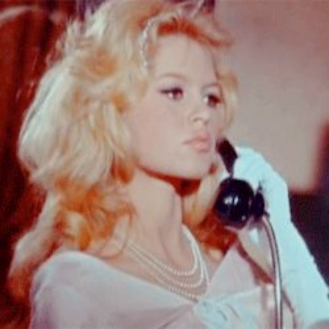 Brigette Bardot is in an interior setting, only her head and shoulders shown in the image.  She is standing up, wearing a white, lacy blouse, elbow length white gloves and has several strings of pearls around her neck.  She has a black phone receiver to her ear with a serious look on her face.  Her blond hair is layered and curled and falls around her shoulders.