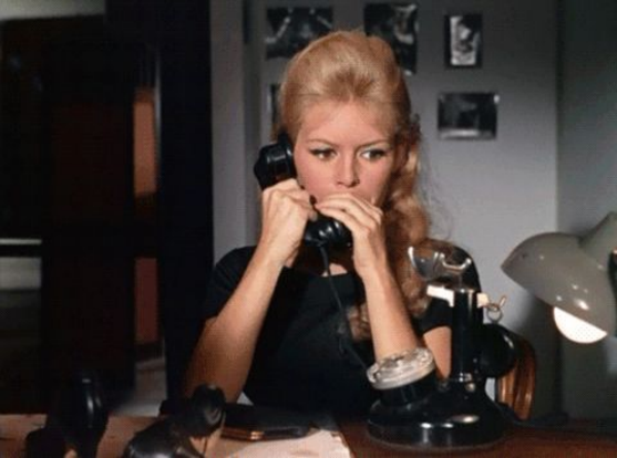 Brigette Bardot is in an office setting in a still image from a movie.  There is a lamp on the desk and the scene is fairly dark.  Brigette is using the phone and has her hand over the mouthpiece and her mouth as if she is being secretive about the call.   She is shown from the waist up, wearing a black shirt, both of her arms are up in front of her.