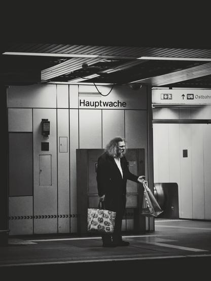 The image is a portrait-orientated black and white photograph of man standing on a subway platform. He has long, curly hair and is wearing a dark suit jacket over a lighter shirt. He's holding two shopping bags, one in each hand. Behind the person is a wall with a sign that reads "Hauptwache". There are also directional signs visible, one pointing to "U6 Ostbahnhof". There is a textured pattern along the edge of the platform.