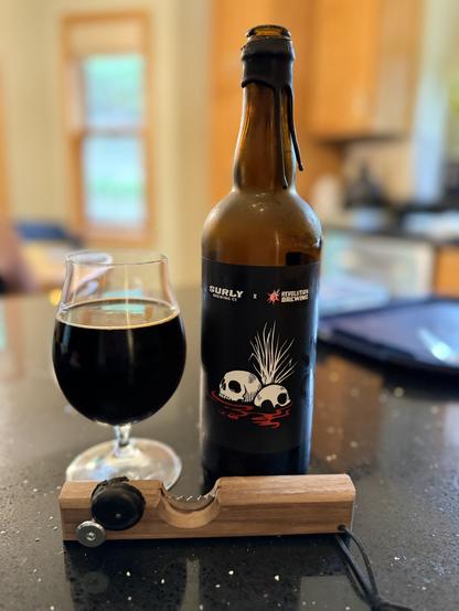 A short snifter glass of dark beer next to a tall brown bottle with a black label. On the label are two skulls and the logos of Surly and Revolution in white and red. A wooden opener is down front.