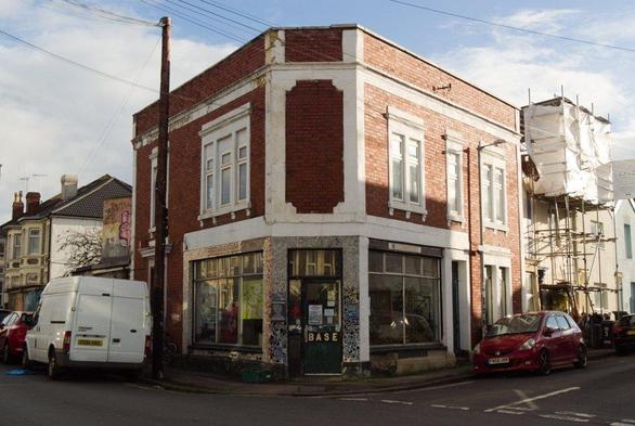 A photo of The Base in Bristol. It is a red brick building with white features. It is shown at the junction of two roads, so has a wedge shaped appearance. There are large shop front windows and an glass entrance door at the front of the wedge.