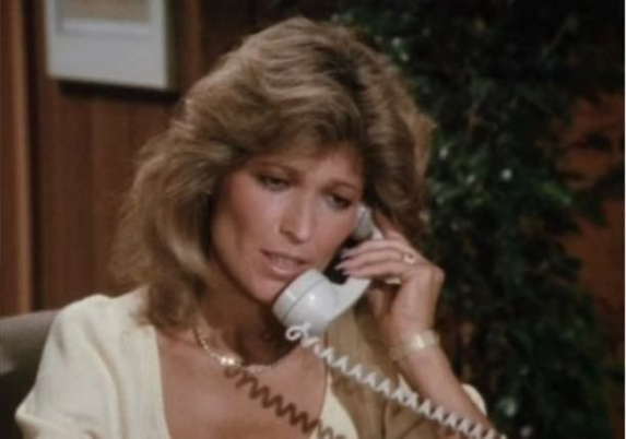 Sherril Lynn Rettino is shown in her role on the TV show "Dallas."  She is on the phone in an office setting, a white receiver in her hand. She is wearing a white blouse.  She seems deep in conversation.  Her feathered hair hangs down to her shoulders.