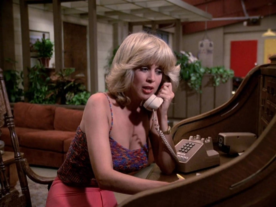 Phyllis Davis in a still image from the TV show Vega$.  She is shown from the waist up, sitting at a curved desk, perhaps in a hotel room or lobby.  She is wearing a red low cut thin strapped blouse and pink shorts.  She is talking on a touch tone phone, which is visible on the desk.  Her blond hair has a flip style and comes down to her shoulders.