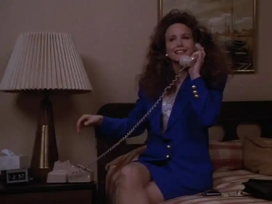 In a still image from the TV show "Murder, She Wrote," Molly Hagen is perched on the side of a bed in what looks like a hotel room.  There is a bedside table with a lamp and the base of the phone on it.  Molly has the phone receiver up to her ear and looks cheerful.  She is wearing a dark blue jacket and matching skirt, a white blouse is below the jacket.  Her legs are crossed and she is looking in the direction of the camera.  Her long dark hair is very curly and comes down below her shoulder.