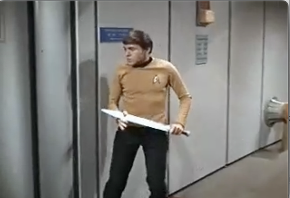 Stillframe from the Star Trek: Original Series episode Day of the Dove. Chekov stands against a corridor wall, a short sword in his hand and look of determination on his face.