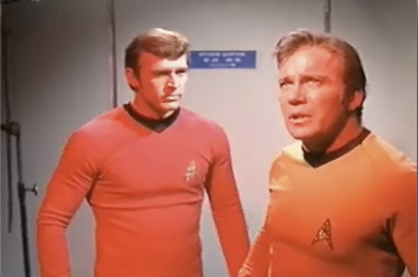 Still frame from Star Trek Original Series episode Day of the Dove. Captain Kirk (right) stands with a lieutenant while a reddish glow covers them. Kirk looks intently towards the ceiling where an off-shot entity spins and whirls away. Watching them.