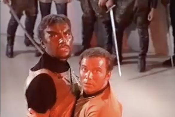 Still frame from Star Trek Original Series episode Day of the Dove. Captain Kirk and Klingon commander Klang hold each other, looking stunned at the whirling, spinning entity off-shot. Their fight interrupted by the entity. A number of Klingon warriors watch on in the far background.