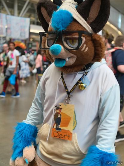 A brown cat-deer fursuiter with blue hair, nose and tongue wearing a cream touque, a pair of oversized glasses, and a white and light blue hoodie, inside the exhibition hall of a convention center.