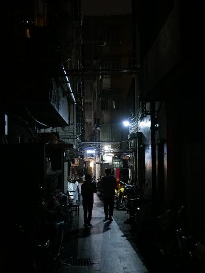 Two people walk down a dark alley. Some lights on either side illuminate their path a little. Surrounding them are buildings and cables