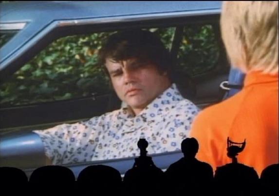 A scene from the film Mitchell, featuring Joe Don Baker in the title role, in a car wearing a floral shirt, displaying a serious expression. Silhouettes of Mystery Science Theater 3000 host Joel Robinson and bots Tom Servo and Crow T. Robot are visible in the foreground, providing their usual comedic riffing.