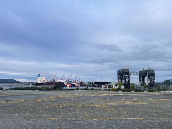 Looking south toward the railyard and barge slip over a wide expanse of old parking lot, riddled with cracks with grass yellowing between them and fading yellow lines on the surface. As a gray cloudy day. There are some large ships in the harbour in the distance and low Islands.