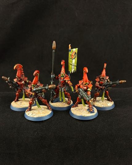 A squad of Warhammer 40K Fire Dragon miniatures painted in shades of deep red with green accents. Each carries a black weapon. The Exarch has a long gun and a back banner.